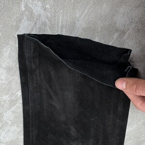 May include: A black suede garment piece featuring a soft, matte exterior texture and a darker, slightly fuzzy interior lining. The material shows visible natural grain variations and a straight stitched seam running vertically along the left side.