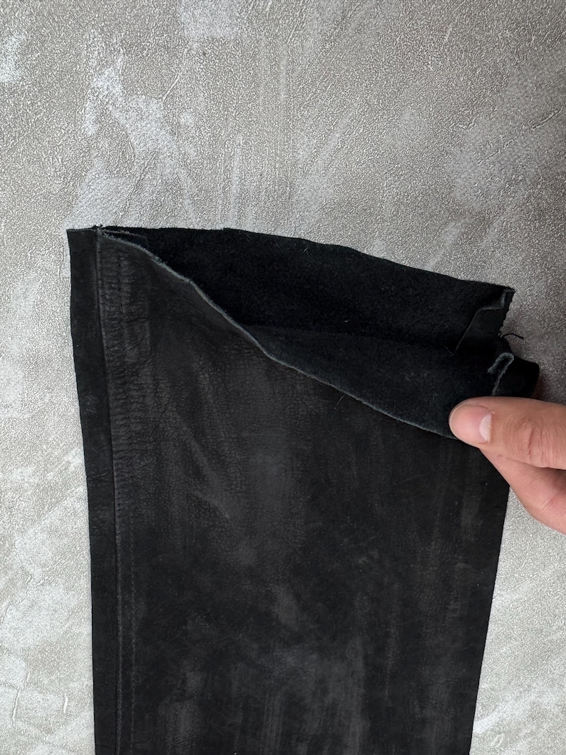 May include: A black suede garment piece featuring a soft, matte exterior texture and a darker, slightly fuzzy interior lining. The material shows visible natural grain variations and a straight stitched seam running vertically along the left side.