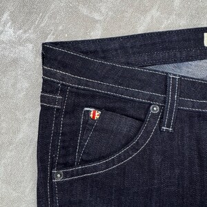 Puede incluir: Pantalones vaqueros de mezclilla azul &iacute;ndigo oscuro con costuras blancas en contraste, dise&ntilde;o cl&aacute;sico de cinco bolsillos y un peque&ntilde;o remache met&aacute;lico circular en el bolsillo monedero con un emblema de la bandera Union Jack en rojo, blanco y azul.