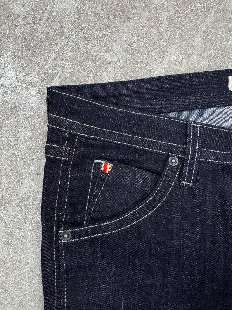 Puede incluir: Pantalones vaqueros de mezclilla azul &iacute;ndigo oscuro con costuras blancas en contraste, dise&ntilde;o cl&aacute;sico de cinco bolsillos y un peque&ntilde;o remache met&aacute;lico circular en el bolsillo monedero con un emblema de la bandera Union Jack en rojo, blanco y azul.