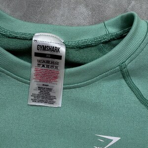 May include: Close-up of a teal Gymshark XS athletic top. The top features a crew neck, long sleeves, and the Gymshark logo in white. A tag with washing instructions is visible. Made of polyester, nylon, and elastane.
