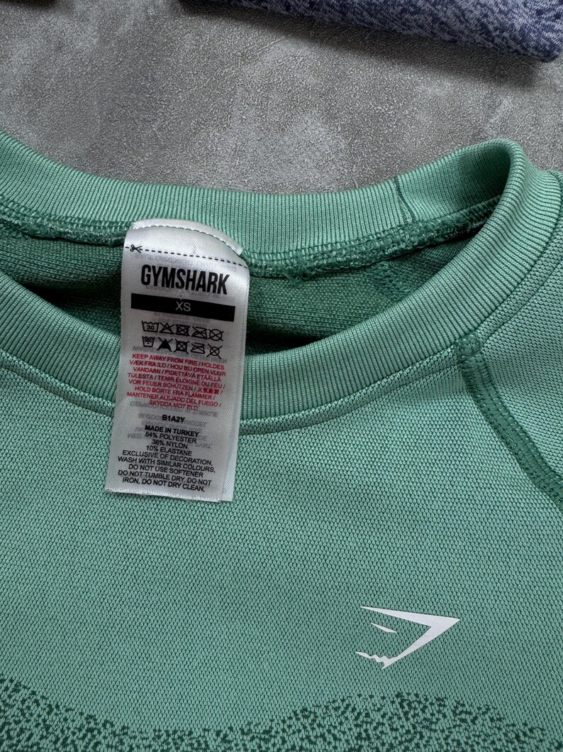 May include: Close-up of a teal Gymshark XS athletic top. The top features a crew neck, long sleeves, and the Gymshark logo in white. A tag with washing instructions is visible. Made of polyester, nylon, and elastane.