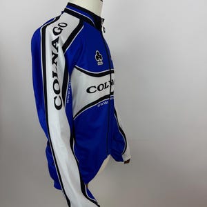 May include: A blue, white, and black Colnago cycling jacket. The jacket has a full zipper, long sleeves, and the Colnago logo on the sleeves and chest. The jacket is displayed on a mannequin.