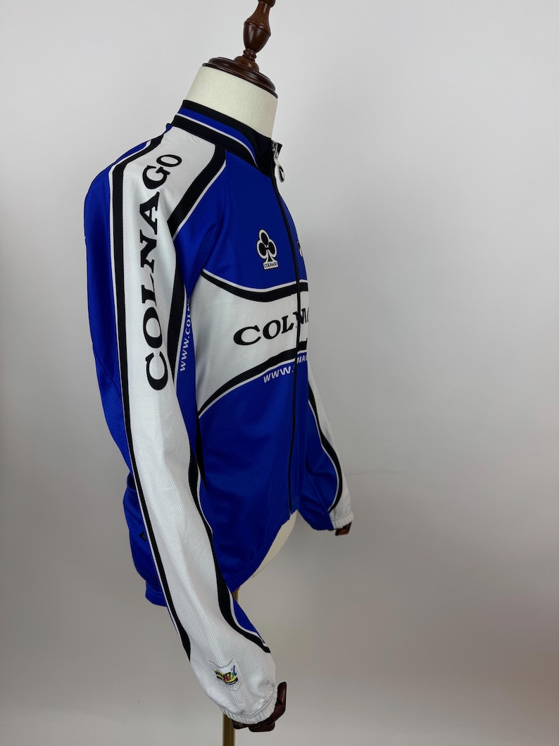 May include: A blue, white, and black Colnago cycling jacket. The jacket has a full zipper, long sleeves, and the Colnago logo on the sleeves and chest. The jacket is displayed on a mannequin.