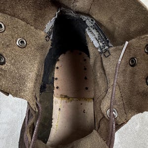May include: An inside view of a brown leather boot. The boot's interior reveals a worn, textured insole and the stitching details. The boot has metal eyelets for laces and a yellow tag with text.