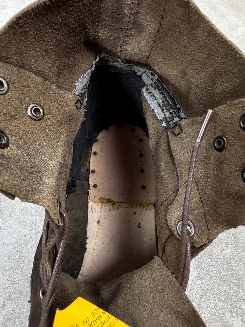 May include: An inside view of a brown leather boot. The boot's interior reveals a worn, textured insole and the stitching details. The boot has metal eyelets for laces and a yellow tag with text.