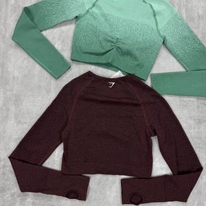 May include: Two long-sleeved athletic tops. One is a deep burgundy color, and the other is a green ombre. Both tops have a textured knit design and thumb holes. The green top has a ruched detail at the waist.