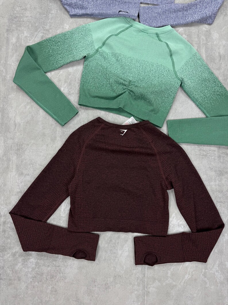 May include: Two long-sleeved athletic tops. One is a deep burgundy color, and the other is a green ombre. Both tops have a textured knit design and thumb holes. The green top has a ruched detail at the waist.