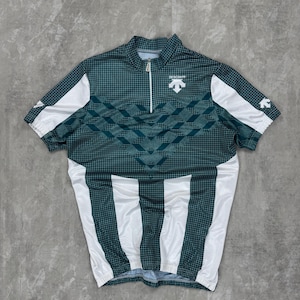 Op de afbeelding: Een korte mouwen wielertrui in teal en wit. De trui heeft een geometrisch patroon op de bovenborst en een verticaal streepontwerp op de onderkant. De trui heeft een rits en de merknaam "DESCENTE" is zichtbaar.