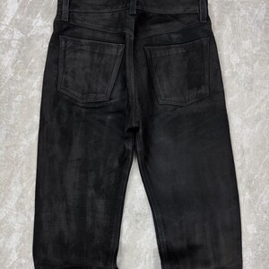May include: A pair of black leather trousers shown from the back, featuring a waistband with belt loops, a central vertical seam, and two square-shaped patch pockets on the rear. The material has a matte, slightly textured finish with tonal variations.