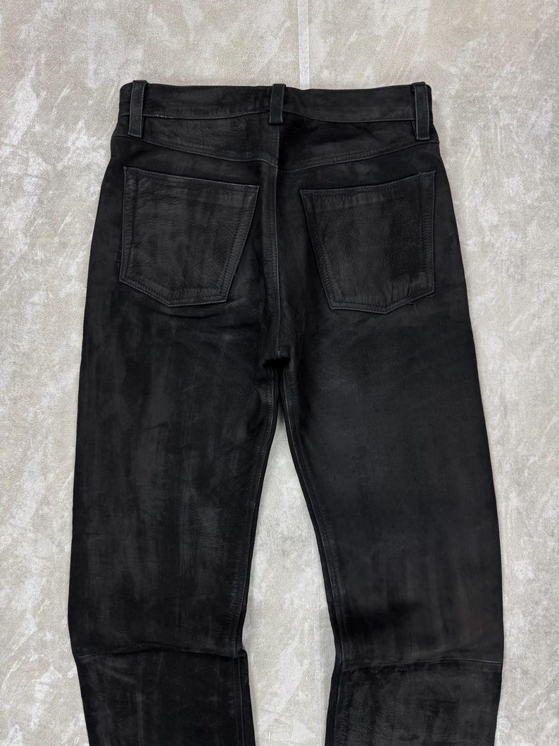 May include: A pair of black leather trousers shown from the back, featuring a waistband with belt loops, a central vertical seam, and two square-shaped patch pockets on the rear. The material has a matte, slightly textured finish with tonal variations.