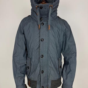 May include: A slate blue jacket with a high collar and a ribbed knit hem and cuffs. The jacket features a button-up front, two front pockets, and a drawstring detail on the collar. The jacket is displayed on a wooden mannequin.