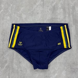 May include: Navy blue swim briefs with yellow and black stripes on the sides. The briefs feature the Olympic rings and the text "OLYMPIC COLLECTION". The H&M logo is visible on the right side.
