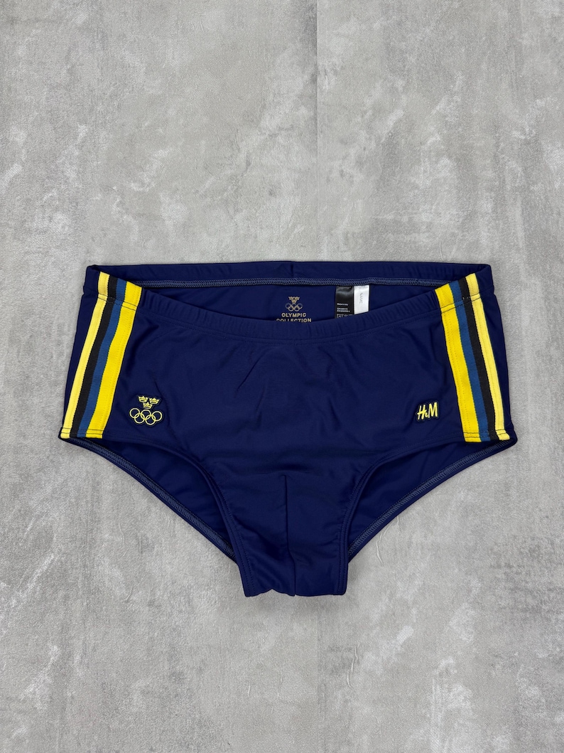 May include: Navy blue swim briefs with yellow and black stripes on the sides. The briefs feature the Olympic rings and the text "OLYMPIC COLLECTION". The H&M logo is visible on the right side.