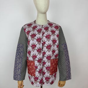 May include: A quilted jacket featuring a floral pattern in pink and red on a white backdrop. The sleeves are grey with a geometric design on the lower sleeves. Two red-brown pockets are on the front.