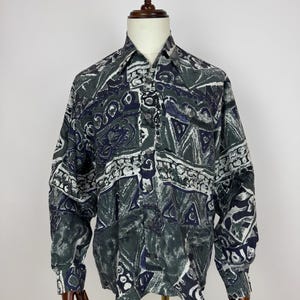 Puede incluir: Camisa de manga larga con un estampado geométrico en tonos verdes, azul marino y blanco. La camisa se exhibe en un maniquí, mostrando su cuello y la parte delantera abotonada. El patrón incluye formas y líneas abstractas.