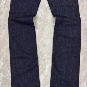 Puede incluir: Un par de pantalones vaqueros de mezclilla en color azul &iacute;ndigo oscuro con corte de pierna recta y costuras tonales visibles en los dobladillos. La tela muestra una textura de tejido sutil y uniforme a lo largo de las piernas, desde el muslo hasta los tobillos.
