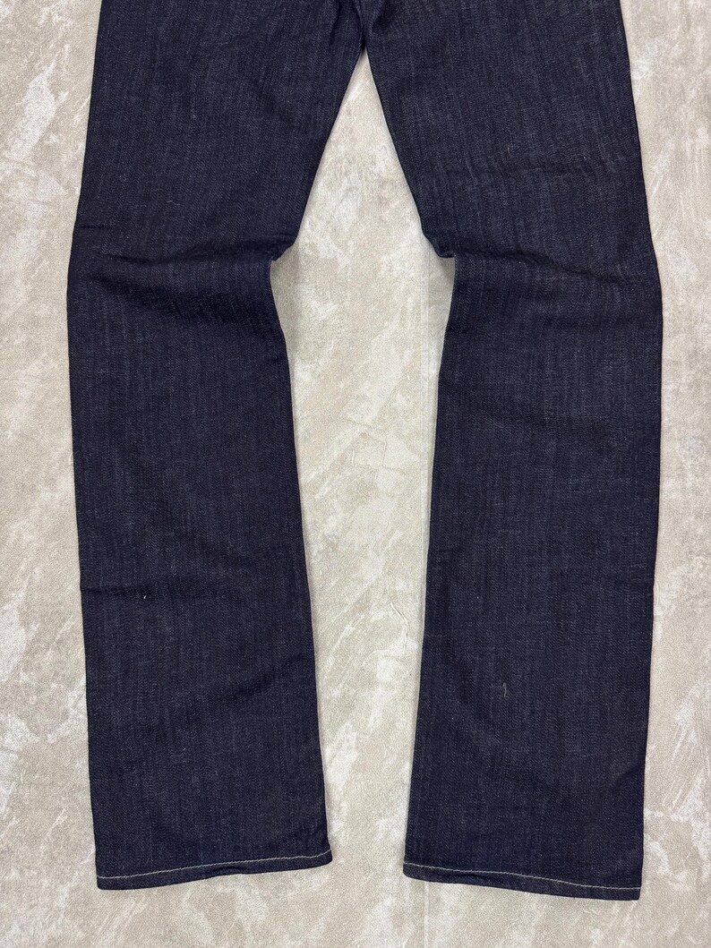 Puede incluir: Un par de pantalones vaqueros de mezclilla en color azul &iacute;ndigo oscuro con corte de pierna recta y costuras tonales visibles en los dobladillos. La tela muestra una textura de tejido sutil y uniforme a lo largo de las piernas, desde el muslo hasta los tobillos.