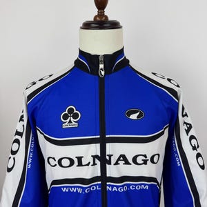May include: Blue, white, and black cycling jacket with the Colnago logo and branding. The jacket has a full-length zipper and a stand-up collar. The sleeves and shoulders are white, with black accents. The front features the Colnago logo and website.