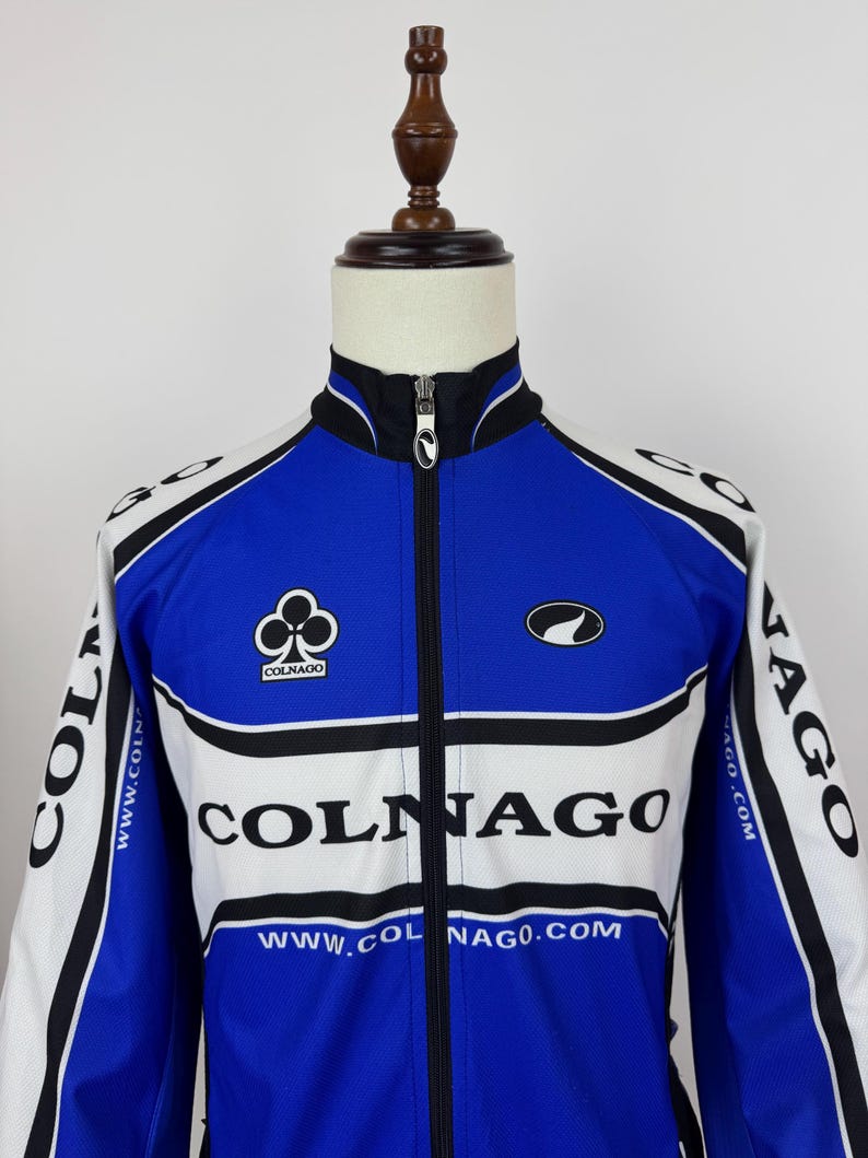 May include: Blue, white, and black cycling jacket with the Colnago logo and branding. The jacket has a full-length zipper and a stand-up collar. The sleeves and shoulders are white, with black accents. The front features the Colnago logo and website.