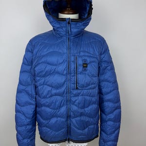 Peut inclure: Veste matelassée bleue avec capuche, fermeture éclair et petite poche poitrine. La veste a des bordures noires aux poignets et à l'ourlet. La veste est présentée sur un mannequin.