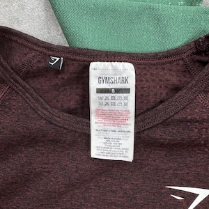 May include: Close-up of a maroon athletic shirt with the Gymshark logo and size tag. The tag includes washing instructions and material composition: 59% nylon, 34% polyester, and 7% elastane. A section of green athletic fabric is visible in the background.