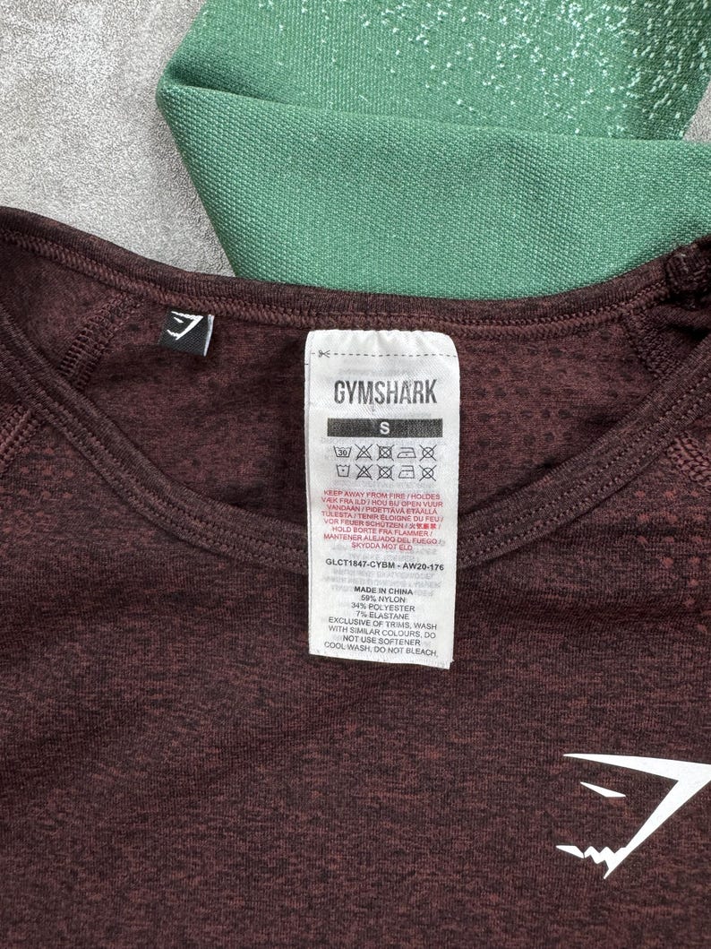 May include: Close-up of a maroon athletic shirt with the Gymshark logo and size tag. The tag includes washing instructions and material composition: 59% nylon, 34% polyester, and 7% elastane. A section of green athletic fabric is visible in the background.