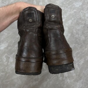 May include: A pair of brown leather boots with the CAT logo embossed on the back. The boots have a worn appearance, with visible wear and tear on the soles and uppers. The boots are a mid-calf height.