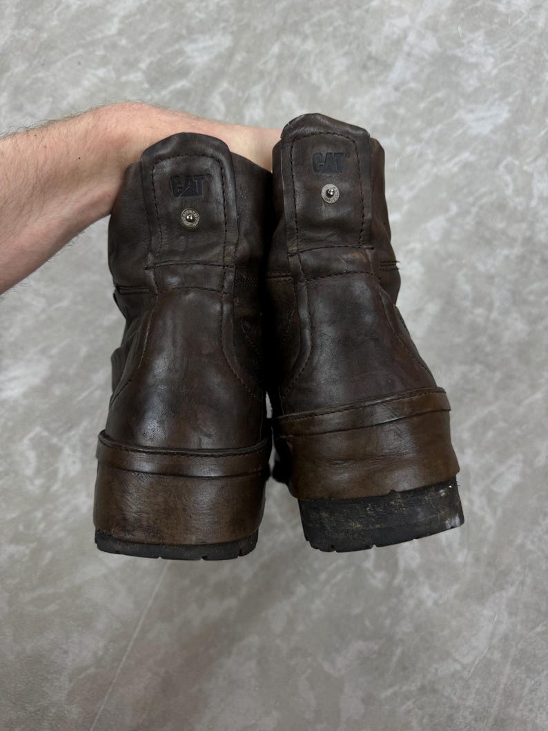 May include: A pair of brown leather boots with the CAT logo embossed on the back. The boots have a worn appearance, with visible wear and tear on the soles and uppers. The boots are a mid-calf height.