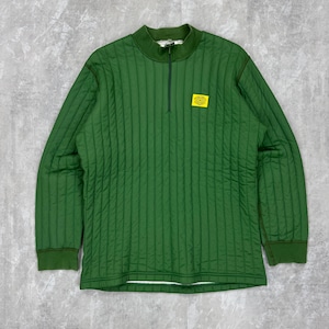 Puede incluir: Una camisa verde acolchada de manga larga con cuello alto y cremallera de cuarto. Tiene un pequeño parche amarillo en el pecho. Las mangas y el cuello son de un verde más oscuro.