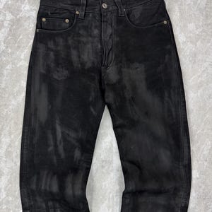 May include: Black denim jeans featuring a distressed, washed-out finish with lighter grey streaks throughout the fabric. The trousers include a standard button fly closure, five pockets, and metallic rivets at the corners of the front pockets.