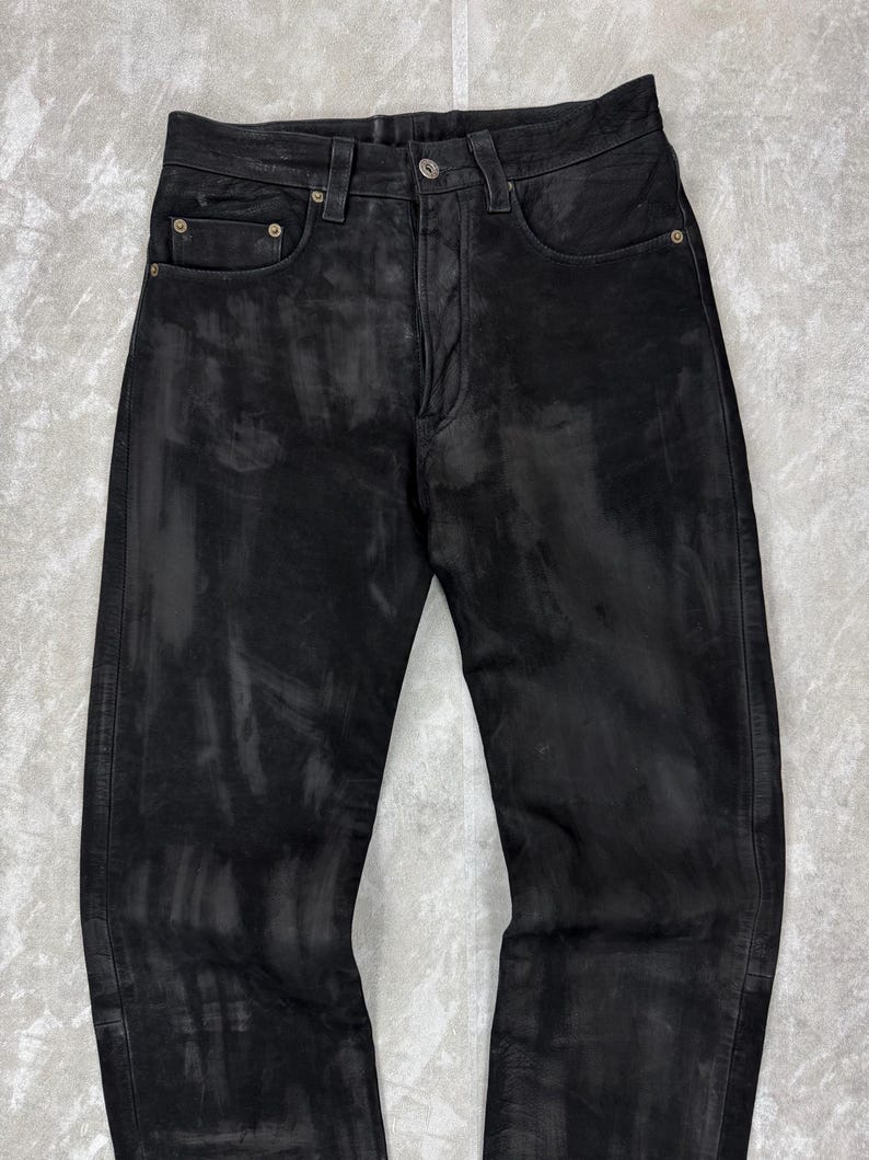 May include: Black denim jeans featuring a distressed, washed-out finish with lighter grey streaks throughout the fabric. The trousers include a standard button fly closure, five pockets, and metallic rivets at the corners of the front pockets.