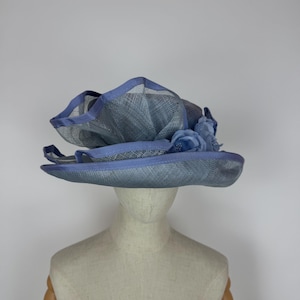 May include: A light blue hat with a wide brim and decorative floral accents. The hat features a layered design with a mesh-like texture and a contrasting darker blue trim. The hat is displayed on a mannequin head.