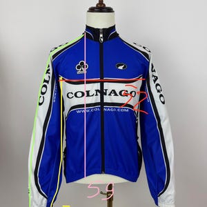 May include: A blue, white, and black Colnago cycling jacket with a full zipper. The jacket features the Colnago logo and website address. The jacket's measurements are 79 inches in length, 52 inches in the chest, and 59 inches in the waist.