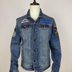 May include: A blue denim jacket with various embroidered patches. The jacket features a button-up front, two chest pockets, and a standard collar. Patches display text such as "SUPERDRY Division" and "ARMY DIVISION".