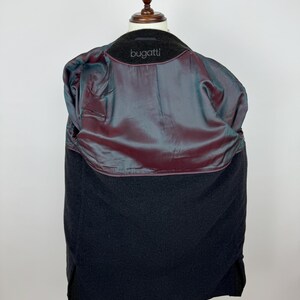 May include: A black wool coat displayed on a wooden mannequin. The coat's interior features a shimmering, iridescent lining in shades of burgundy and teal. The brand name "bugatti" is visible on the collar. The coat is a classic outerwear piece.