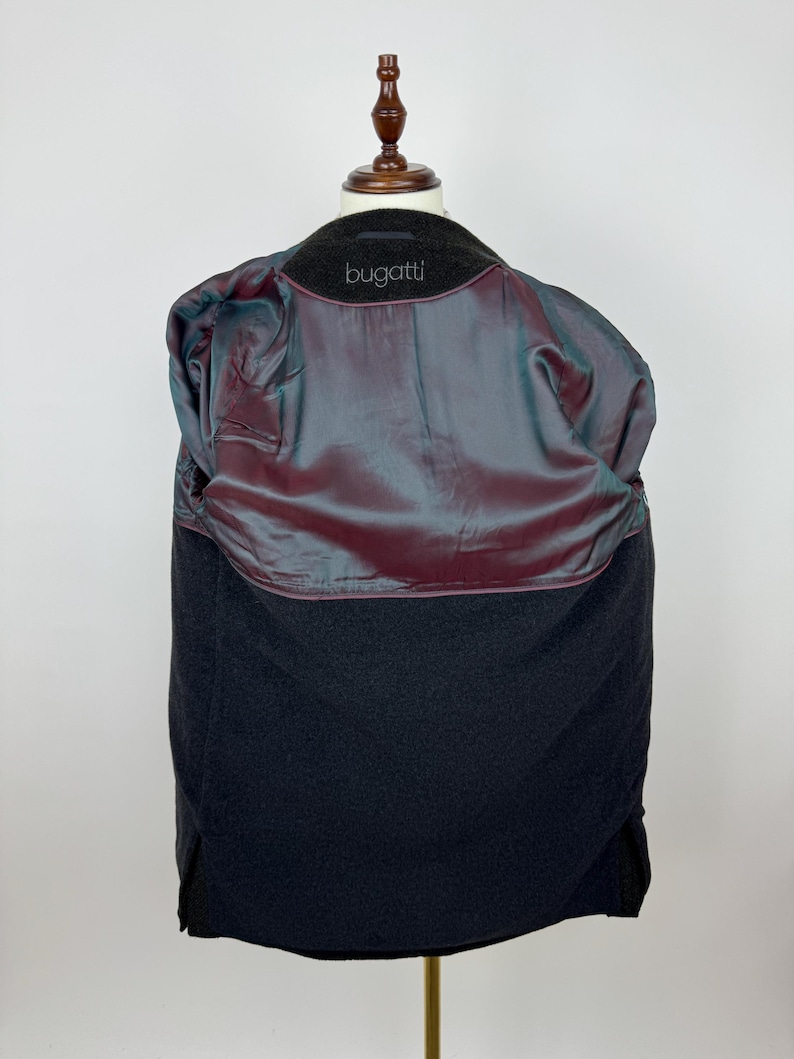 May include: A black wool coat displayed on a wooden mannequin. The coat's interior features a shimmering, iridescent lining in shades of burgundy and teal. The brand name "bugatti" is visible on the collar. The coat is a classic outerwear piece.