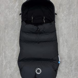 Könnte beinhalten: Schwarzer Baby-Fußsack für Kinderwagen mit Kapuze. Der gesteppte Fußsack hat einen Reißverschluss und ein weißes Logo an der Unterseite. Entwickelt, um ein Baby bei kaltem Wetter warm und bequem zu halten. Misst ca. 90 cm in der Länge.