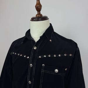 Puede incluir: Camisa negra abotonada con tachuelas plateadas a lo largo del canesú y del bolsillo. La camisa tiene un cuello clásico, un bolsillo en el pecho y costuras blancas. La camisa se exhibe en un maniquí.