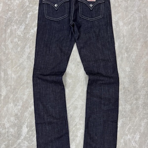 Puede incluir: Pantalones vaqueros de mezclilla en color azul &iacute;ndigo oscuro con dise&ntilde;o cl&aacute;sico de cinco bolsillos. Presentan dos bolsillos traseros con solapa y botones plateados, costuras blancas en contraste y una cintura est&aacute;ndar con trabillas para cintur&oacute;n.