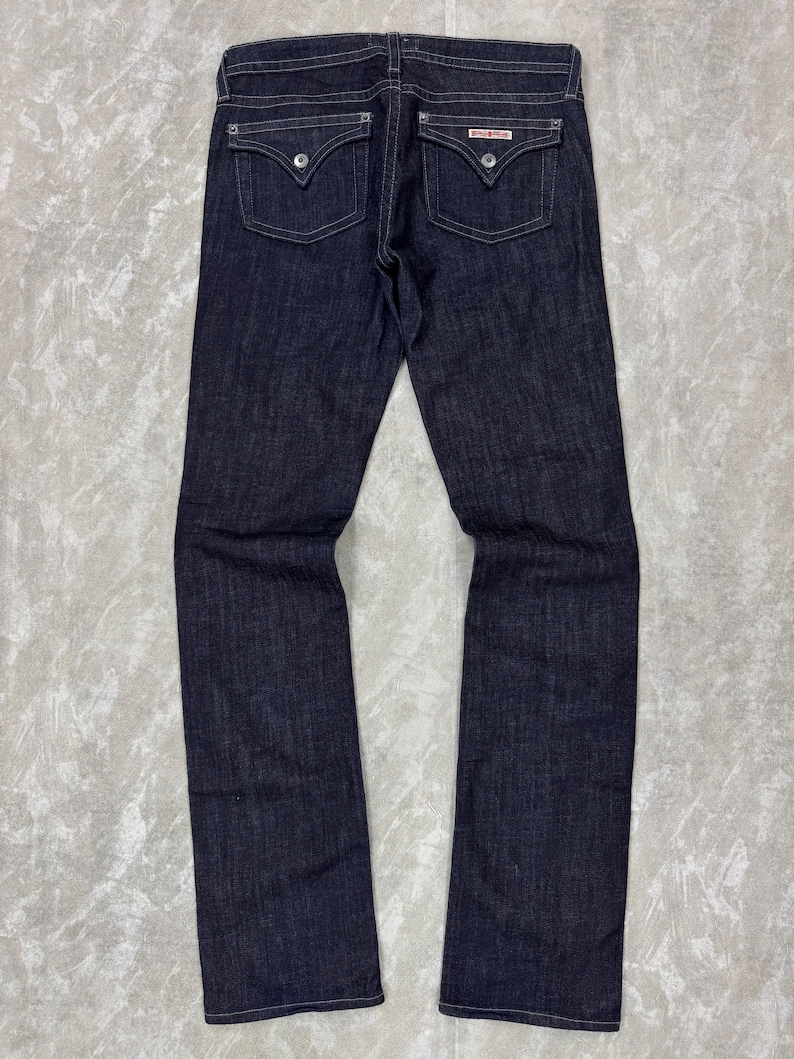 Puede incluir: Pantalones vaqueros de mezclilla en color azul &iacute;ndigo oscuro con dise&ntilde;o cl&aacute;sico de cinco bolsillos. Presentan dos bolsillos traseros con solapa y botones plateados, costuras blancas en contraste y una cintura est&aacute;ndar con trabillas para cintur&oacute;n.