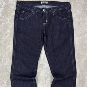 Puede incluir: Pantalones vaqueros de mezclilla en color &iacute;ndigo oscuro con dise&ntilde;o cl&aacute;sico de cinco bolsillos, costuras blancas en contraste, cierre de bot&oacute;n plateado, cremallera y una peque&ntilde;a etiqueta con logo rojo y blanco en el bolsillo delantero derecho.