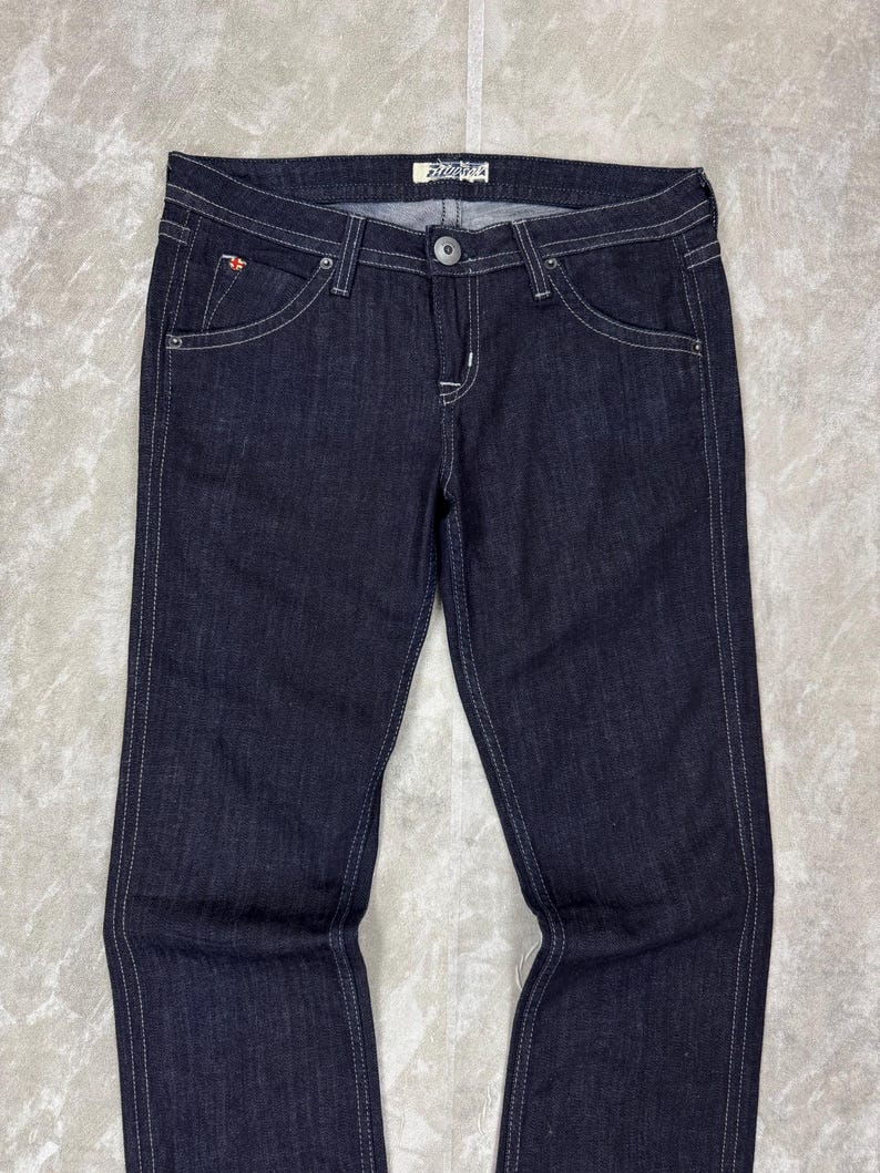 Puede incluir: Pantalones vaqueros de mezclilla en color &iacute;ndigo oscuro con dise&ntilde;o cl&aacute;sico de cinco bolsillos, costuras blancas en contraste, cierre de bot&oacute;n plateado, cremallera y una peque&ntilde;a etiqueta con logo rojo y blanco en el bolsillo delantero derecho.