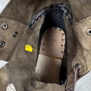 May include: Close-up of the interior of a brown leather boot. The boot features a worn leather insole with perforations, a dark interior, and metal eyelets for laces. A yellow tag with the text "WISAL" is visible.
