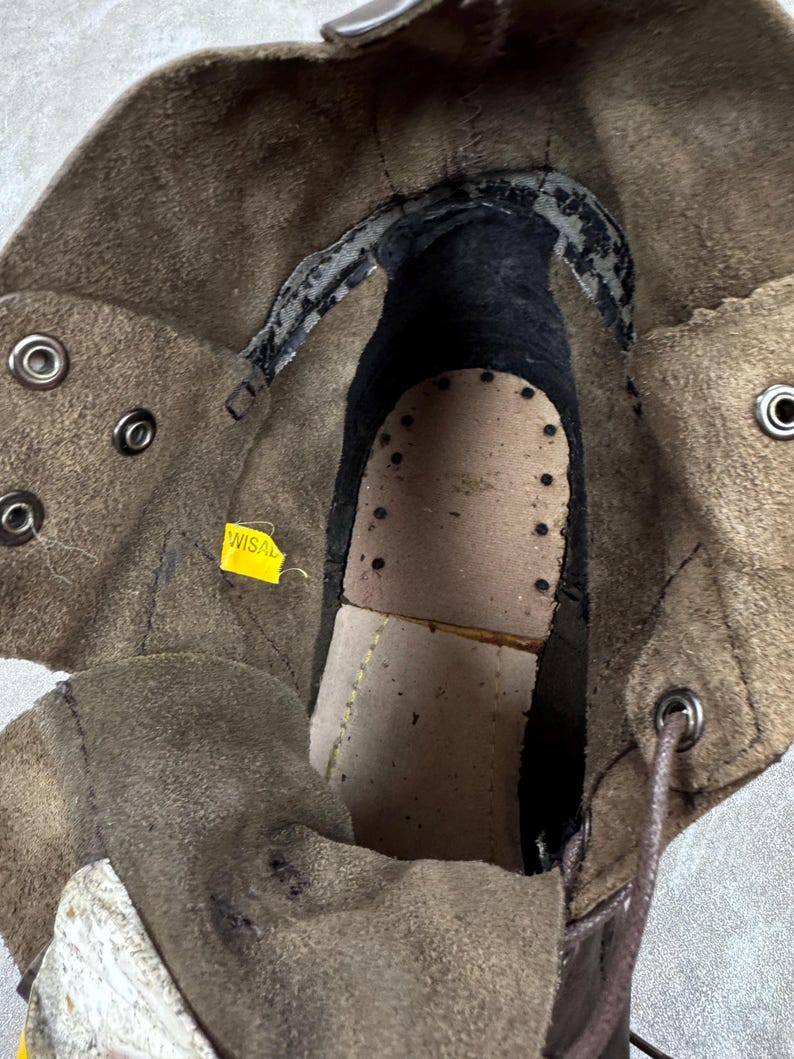 May include: Close-up of the interior of a brown leather boot. The boot features a worn leather insole with perforations, a dark interior, and metal eyelets for laces. A yellow tag with the text "WISAL" is visible.