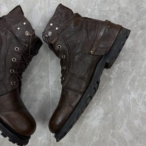 May include: Pair of brown leather boots with a distressed appearance. The boots feature metal eyelets, decorative studs, and a rugged sole. The boots have a lace-up design and a folded-over top.