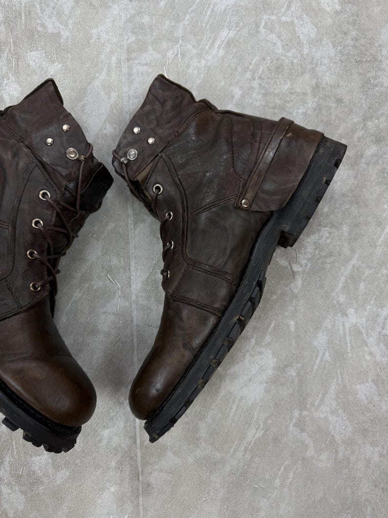 May include: Pair of brown leather boots with a distressed appearance. The boots feature metal eyelets, decorative studs, and a rugged sole. The boots have a lace-up design and a folded-over top.
