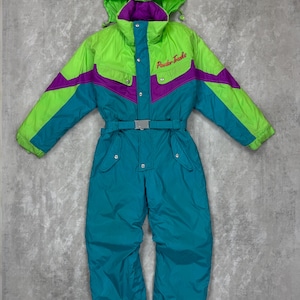 Op de afbeelding: Een vintage skipak in heldere kleuren. Het bovenste deel is limoengroen met paarse en turquoise accenten, en het onderste deel is turquoise. De capuchon is limoengroen en het pak heeft een riem en de woorden "Powder Tracks" op de borst geborduurd.