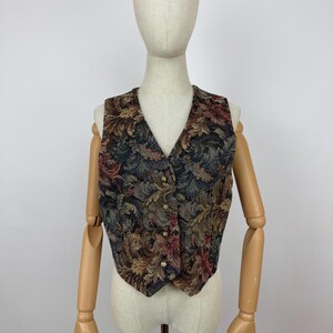 Puede incluir: Un chaleco vintage con cuello en V y cierre de botones. El chaleco presenta un fondo oscuro con un estampado floral en tonos marrones, rojos y dorados. La prenda se exhibe en un maniquí.