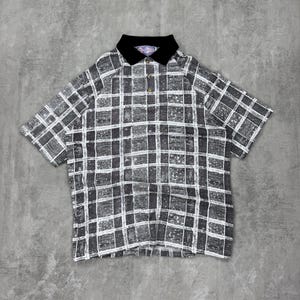 May include: A short-sleeved polo shirt with a black collar and a button-up front. The shirt features a gray and white checkered pattern. The shirt is laid flat on a gray surface.